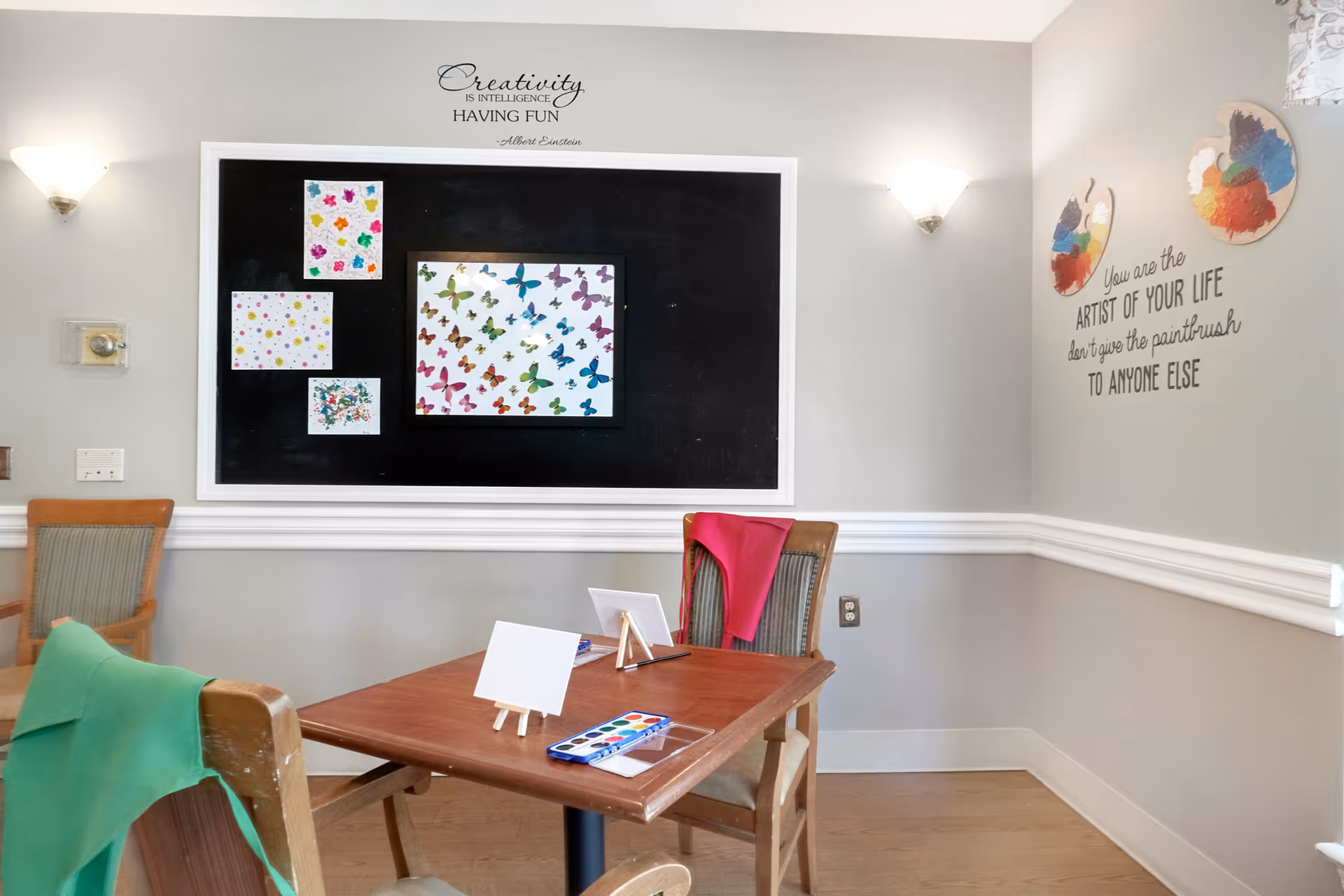 A small art activity room with a wooden table and chairs. On the table are small blank canvases on easels and a watercolor paint set. The walls are decorated with colorful butterfly artwork and inspirational quotes about creativity and being the artist of your life. The room has soft lighting from wall sconces.