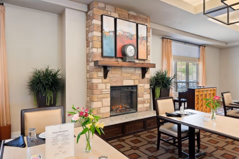 A cozy dining area with tables and chairs, featuring a stone fireplace with a clock and three-panel artwork above it. The room has large windows with peach-colored curtains, potted plants on either side of the fireplace, and a piano in the background. Tables are set with menus, glasses, and napkins, and there are flower vases on the tables.