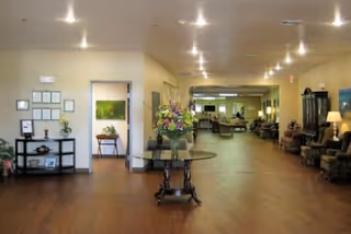 Spacious, well-lit senior living facility interior with a central table of flowers, seating along the sides, and a long hallway leading to more common areas.