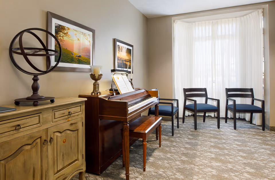 A cozy room with a wooden upright piano and matching bench, three dark wooden chairs with blue cushions arranged near a large window with sheer white curtains. A rustic wooden cabinet with decorative items and framed landscape photographs hang on the beige walls.