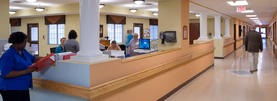 Interior view of a nursing home reception desk and adjoining corridor with staff working at the desk and a resident walking down the hallway.