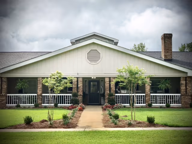 Front exterior view of a single-story building with a gabled roof, brick walls, white railings, and a central entrance door. The building is surrounded by a well-maintained lawn with small trees and shrubs along a concrete walkway leading to the entrance. The sky is cloudy.
