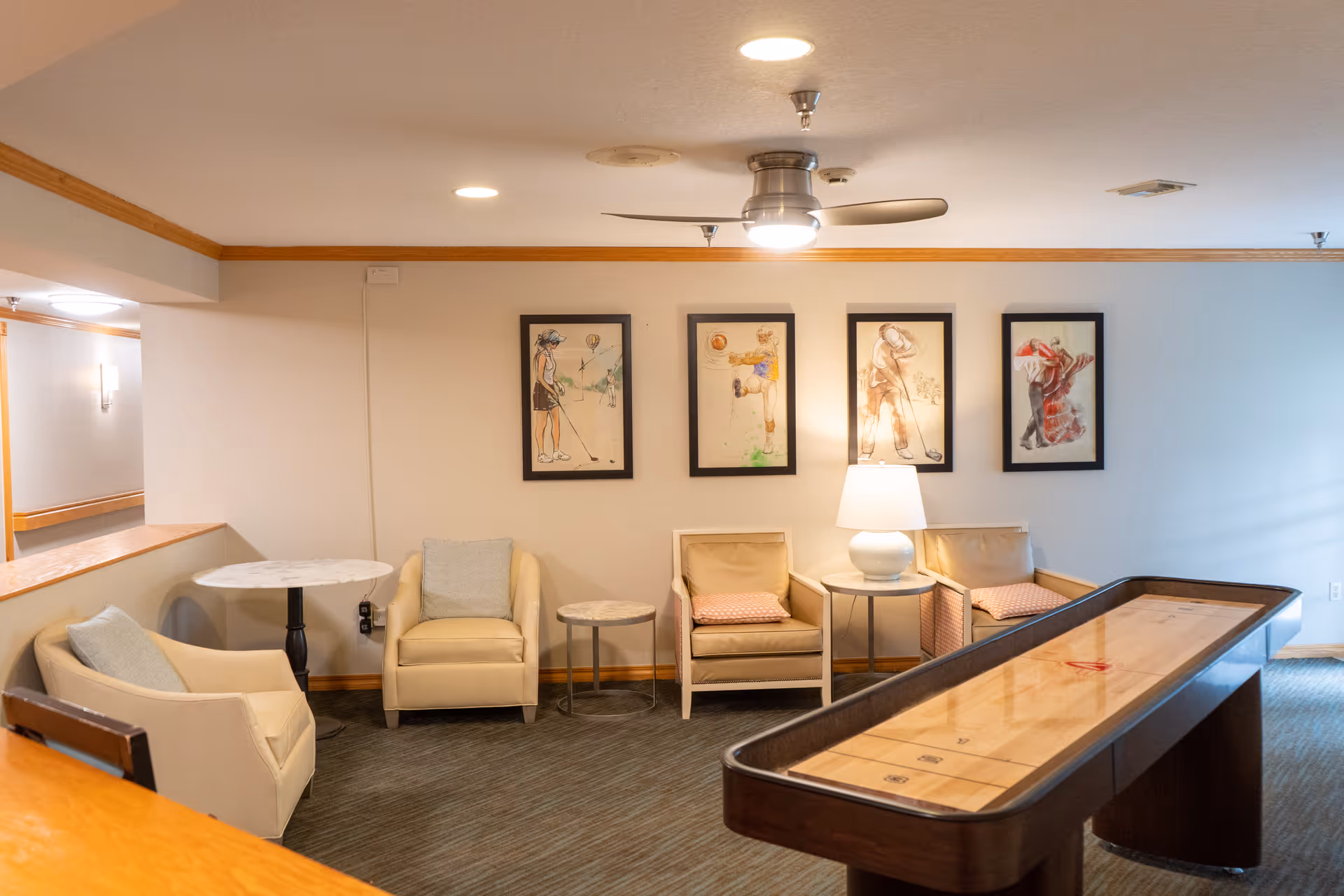 A cozy common area with four armchairs arranged along a wall, each with a cushion. There are three small round tables, one with a lamp. Above the chairs, four framed artworks depict people playing various sports. A ceiling fan with a light is mounted on the ceiling. In the foreground, there is a shuffleboard table.