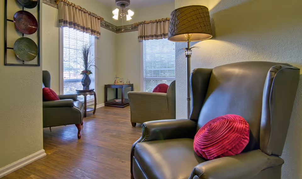 A cozy sitting area with two leather armchairs, each with a red round pillow. There is a floor lamp with a beige lampshade next to one chair. The room has wooden flooring, two large windows with blinds and valances, and a small table with a vase of flowers between the windows. Decorative wall art with colorful plates is mounted on the left wall.
