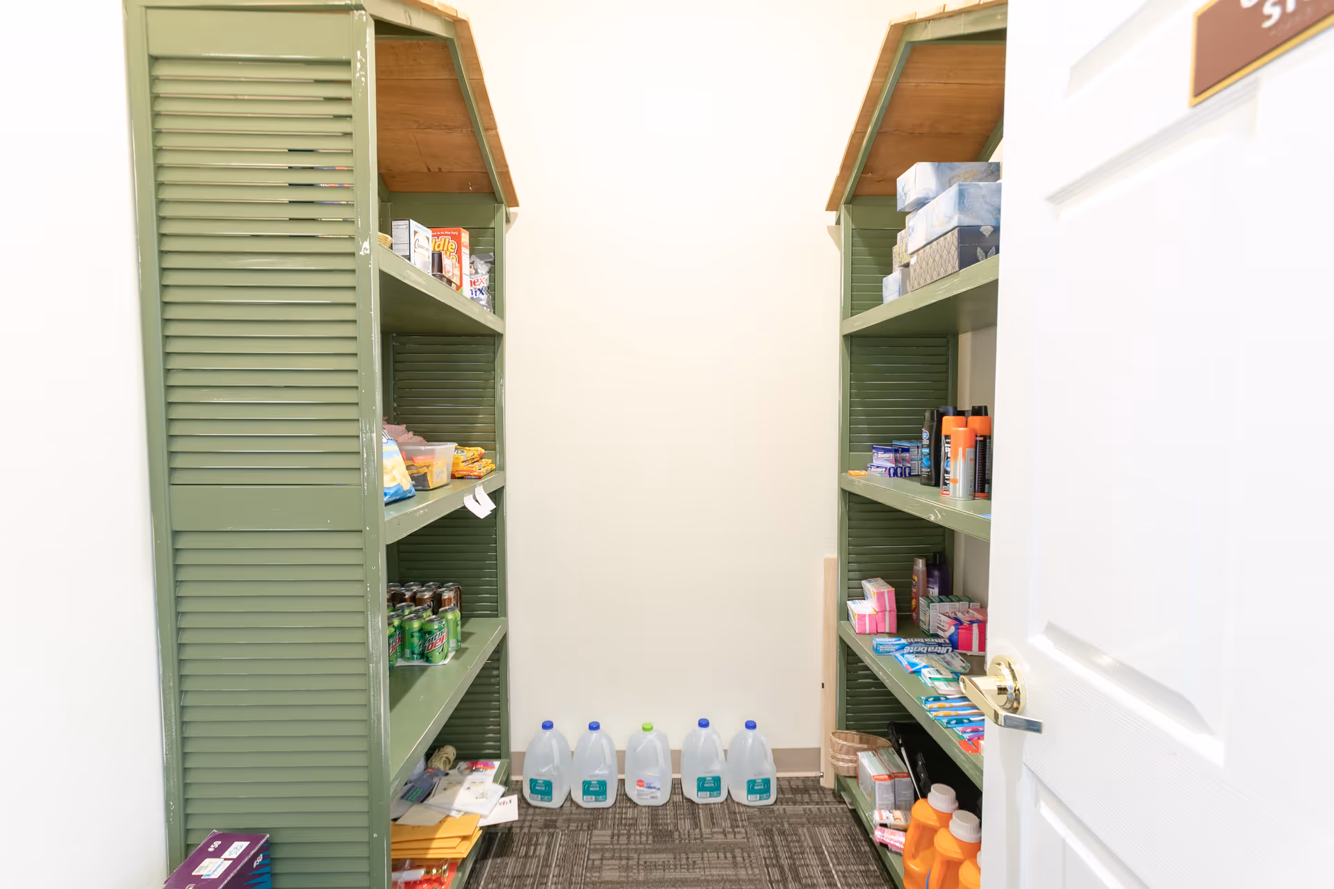 A small storage pantry with green shelves on both sides stocked with various items including canned drinks, boxes, tissue boxes, and cleaning supplies. Five gallon water jugs are lined up on the floor at the back wall. The door to the pantry is partially open.