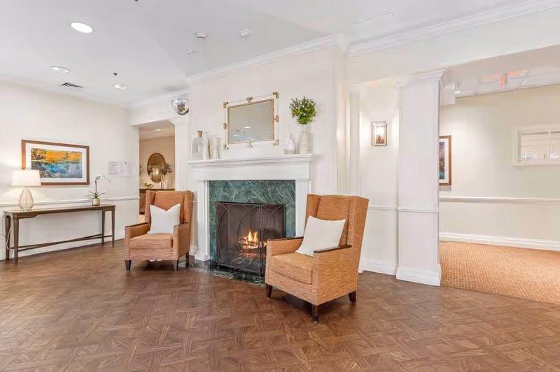 A cozy interior common area featuring a lit fireplace with a green marble surround, two orange armchairs with white pillows placed on either side of the fireplace, a wooden console table with a lamp and a framed painting on the wall, and neutral-colored walls and flooring.