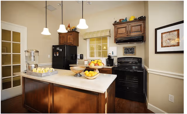 A kitchen area with wooden cabinets, a black refrigerator, and a black stove with a range hood. There is a kitchen island with a countertop holding a water dispenser, a tray with cups, and bowls of fruit. Three pendant lights hang from the ceiling. A framed coffee-themed picture is on the wall to the right.