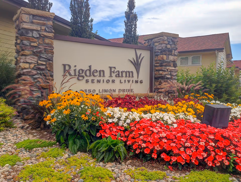 Entrance sign for Rigden Farm Senior Living surrounded by colorful flower beds with red, white, yellow, and orange flowers, stone pillars on either side of the sign, and a building with a red roof and beige siding in the background under a partly cloudy sky.