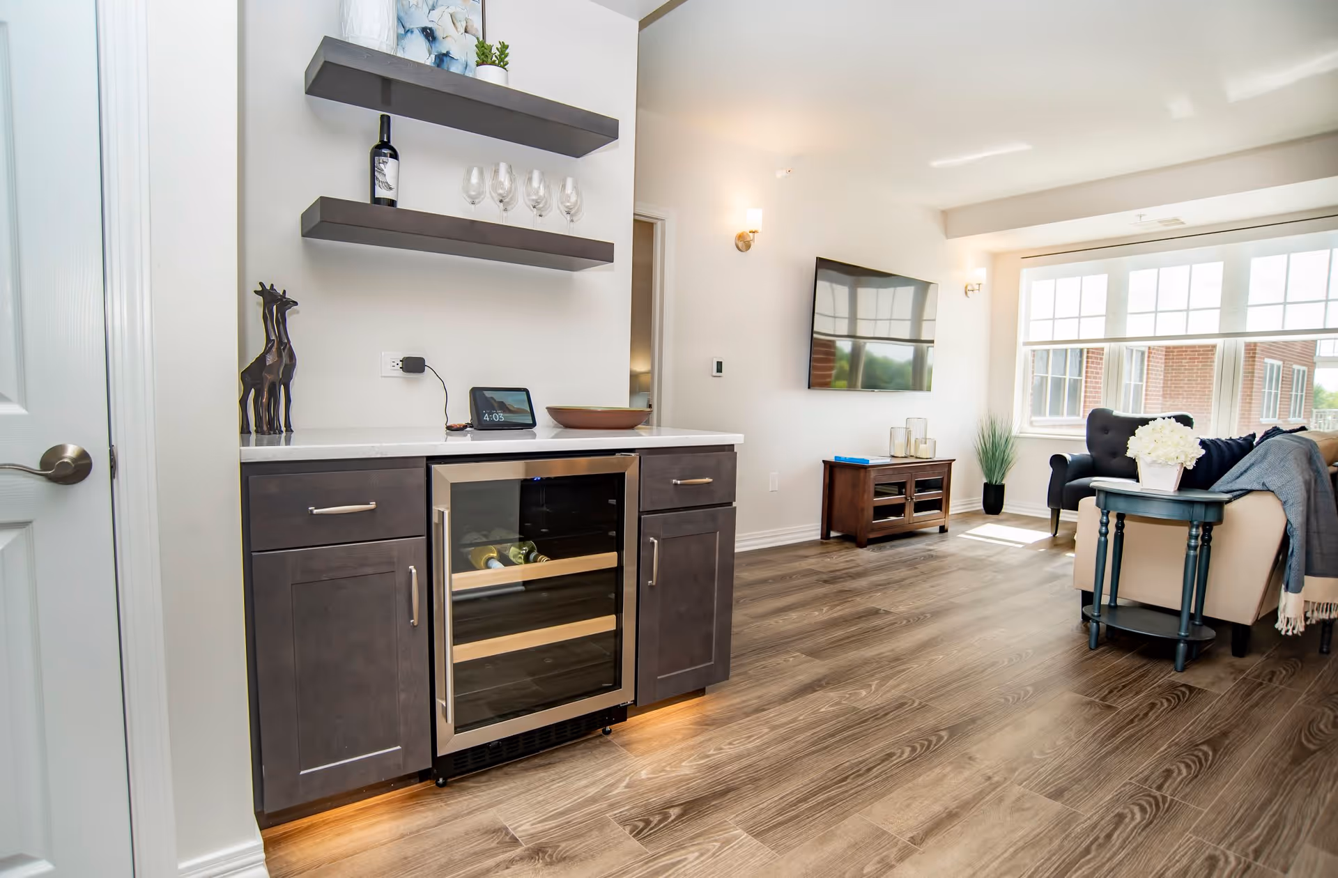 A bright and modern living room area with wood flooring, a mounted flat-screen TV, a small dark wooden cabinet, and a seating area with a beige sofa and a black armchair. There is a small table with a white flower arrangement next to the sofa. On the left side, there is a built-in cabinet with a wine cooler, two floating shelves holding wine glasses, a bottle of wine, and decorative items. Large windows allow natural light to fill the room.