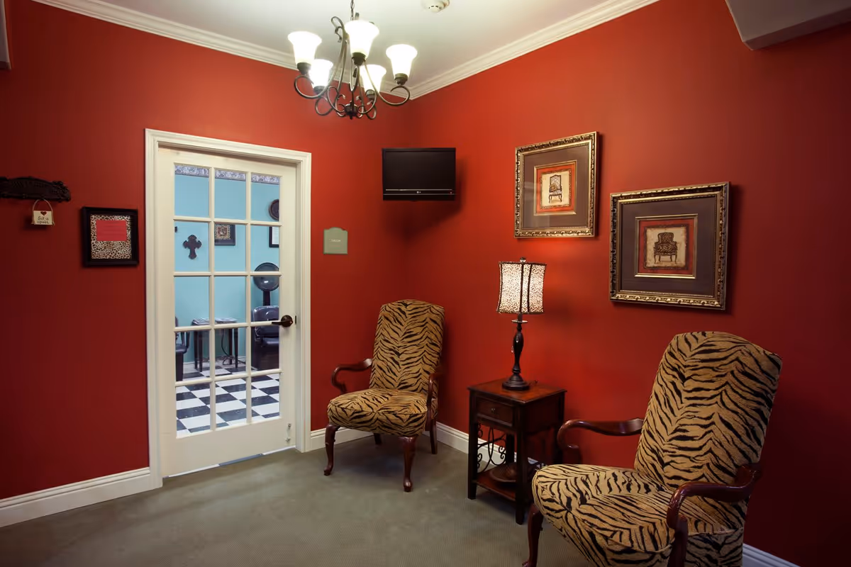 A cozy sitting area with two tiger-striped upholstered armchairs facing each other, a small wooden side table with a lamp between them, and two framed pictures of chairs on the red wall behind. A chandelier hangs from the ceiling, and a glass-paneled door leads to another room with black and white checkered flooring and teal walls.