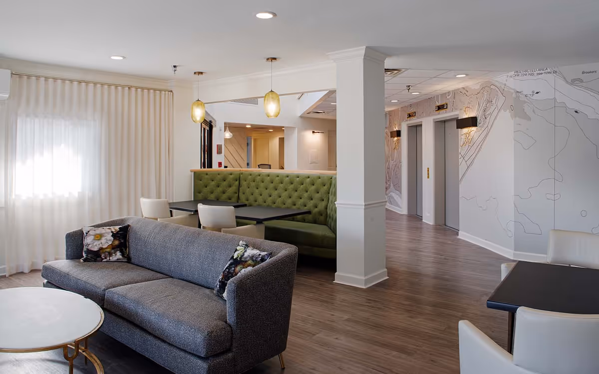A modern and cozy common area in a senior living facility featuring a gray sofa with floral pillows, a round white coffee table, green tufted banquette seating with black tables, white chairs, pendant lights, and two elevators in the background with a wall mural.