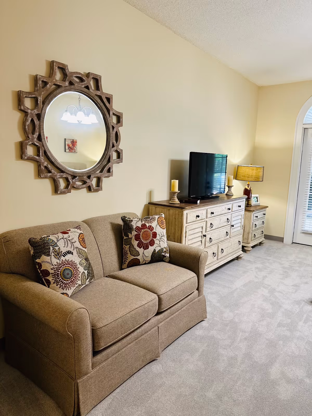 A cozy living room area with a beige loveseat adorned with two floral patterned pillows. Above the loveseat is a decorative round mirror with an intricate wooden frame. Across from the loveseat is a wooden dresser with a flat-screen TV on top, flanked by two candle holders and a table lamp. The room has light-colored walls and carpeted flooring, with a door featuring a large window and blinds in the background.