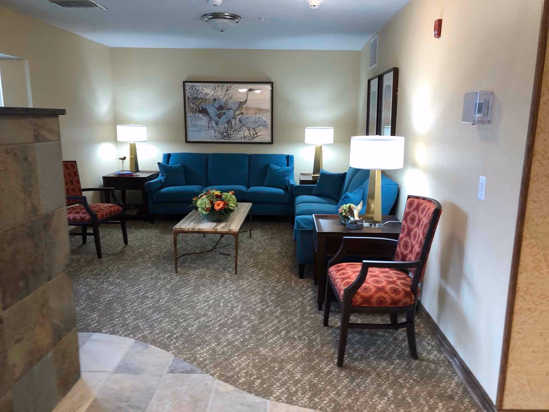 A cozy living room area with a blue sofa and matching loveseat, two red patterned armchairs, a wooden coffee table with a floral centerpiece, two side tables with lamps, and a framed artwork of birds on the wall.