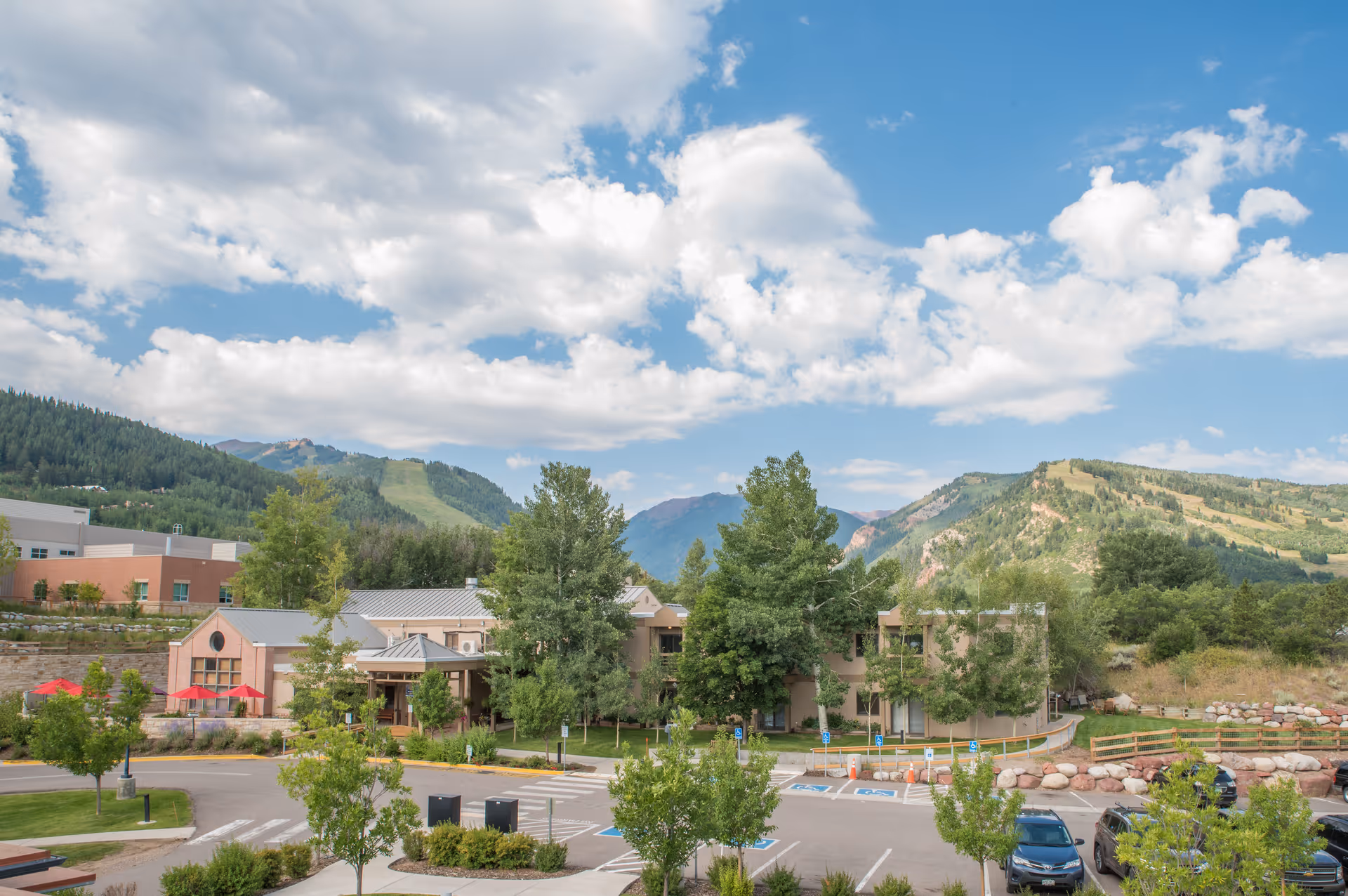 View of Aspen Valley Health – Whitcomb Terrace facility surrounded by trees and greenery with mountains in the background under a partly cloudy blue sky. There is a parking lot with several cars and handicap parking spaces in front of the building.