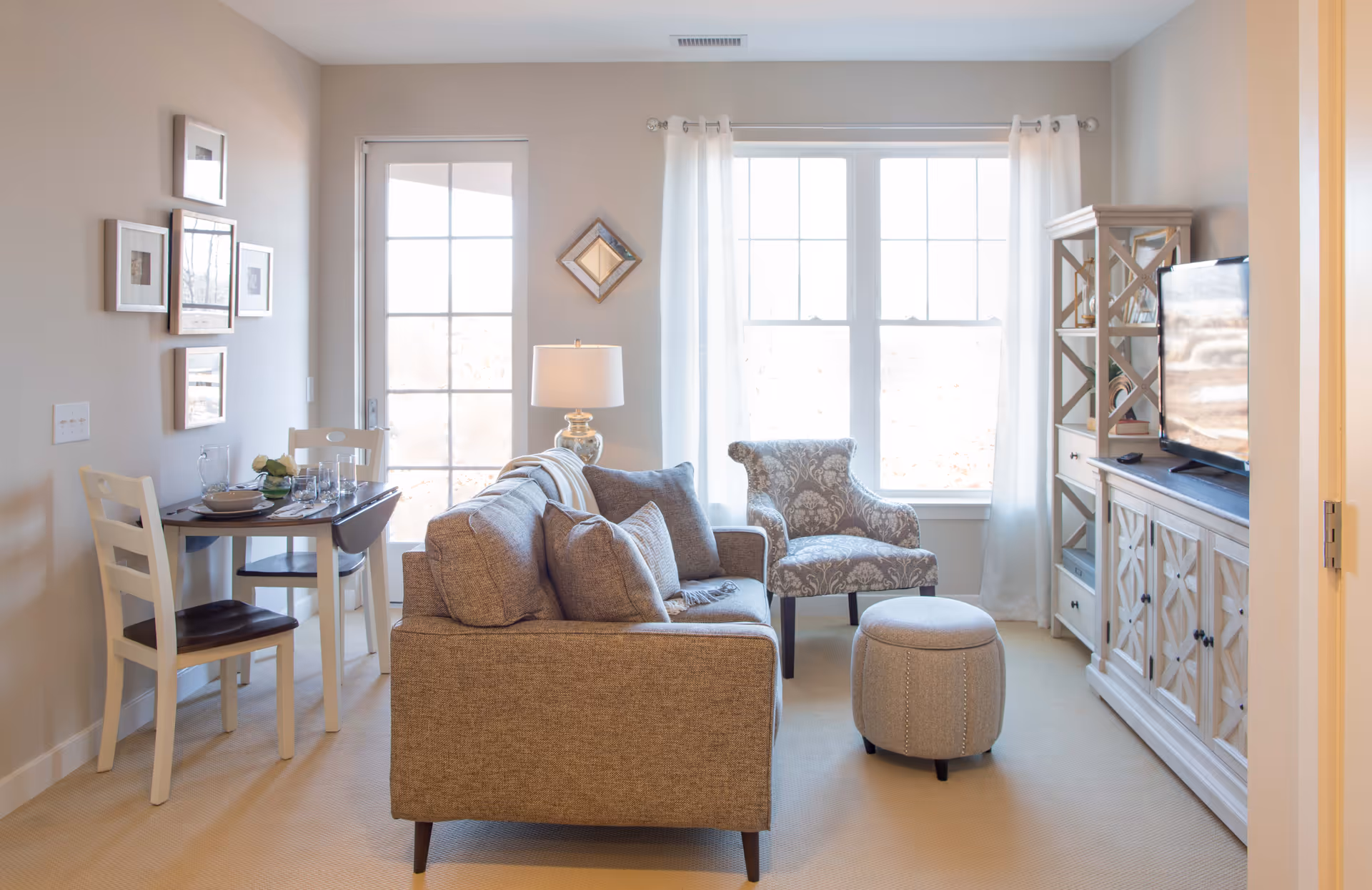 Sunlit living room with a sofa, patterned armchair and ottoman, TV on a console, and a small dining table by a window and glass door.