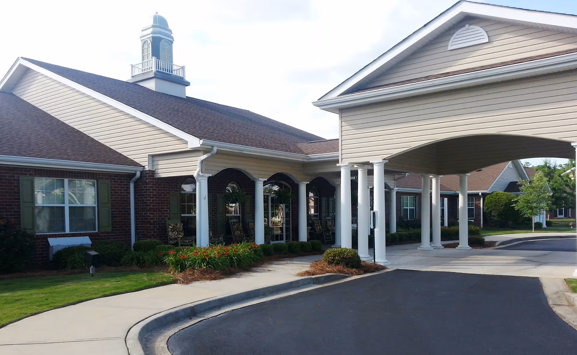 Exterior view of a senior living facility with a covered entrance supported by white columns, brick walls, green window shutters, and a small landscaped area with bushes and flowers.