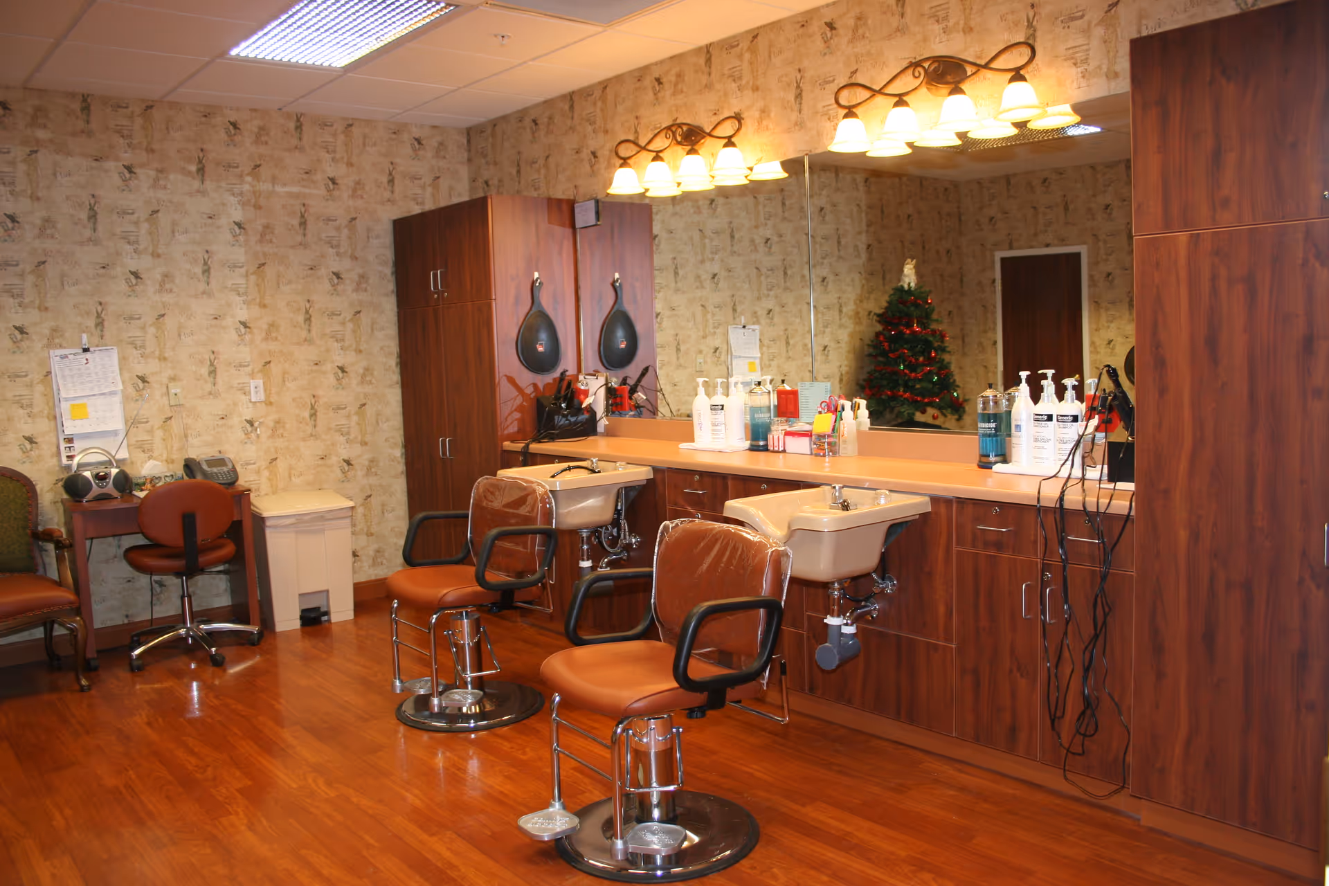 Interior view of a salon area in a nursing center with two brown salon chairs in front of sinks and a large mirror. The room has wooden flooring, wood cabinetry, and wallpapered walls. There is a small decorated Christmas tree reflected in the mirror, a desk with a chair, a phone, and a radio on the left side.