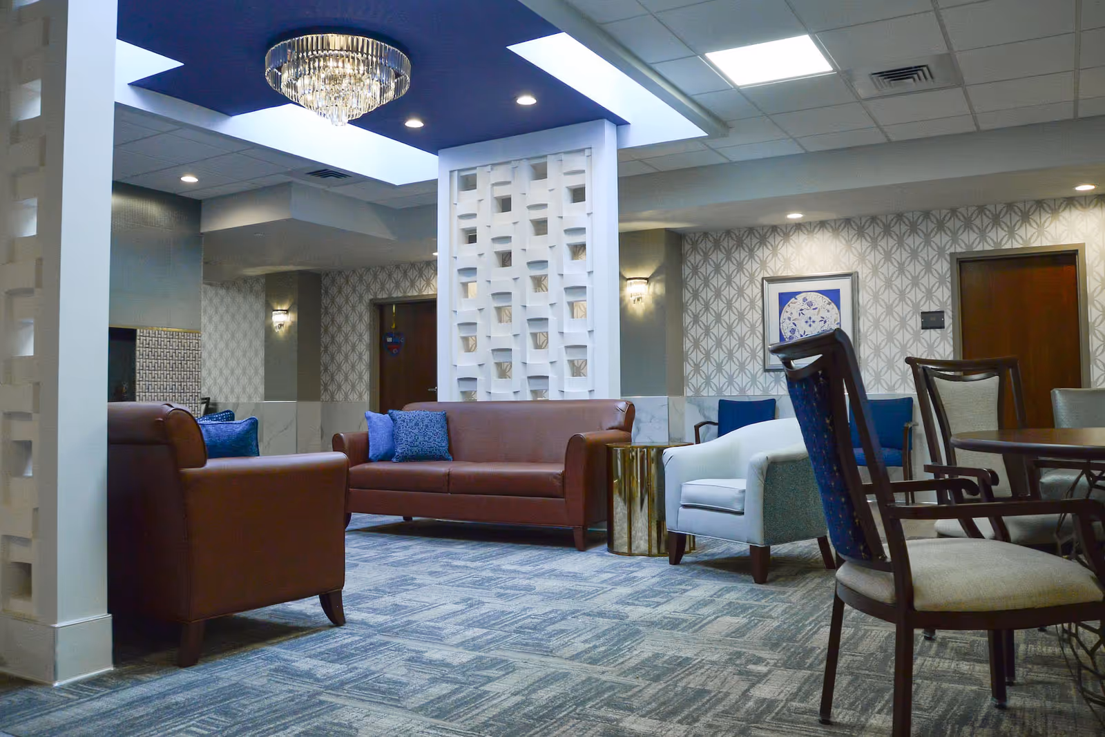 A well-lit lounge area with brown leather sofas, a white armchair, and several wooden chairs around a table. The room features patterned wallpaper, a decorative white partition, a chandelier, and a carpeted floor.