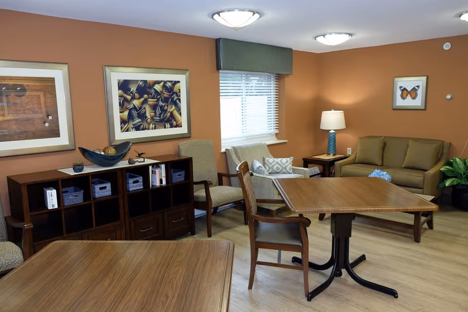 A cozy living room with orange walls featuring a wooden table with chairs, a beige sofa, two armchairs, a wooden cabinet with decorative items and books, framed artwork on the walls, a window with blinds and a green valance, and a table lamp on a side table.