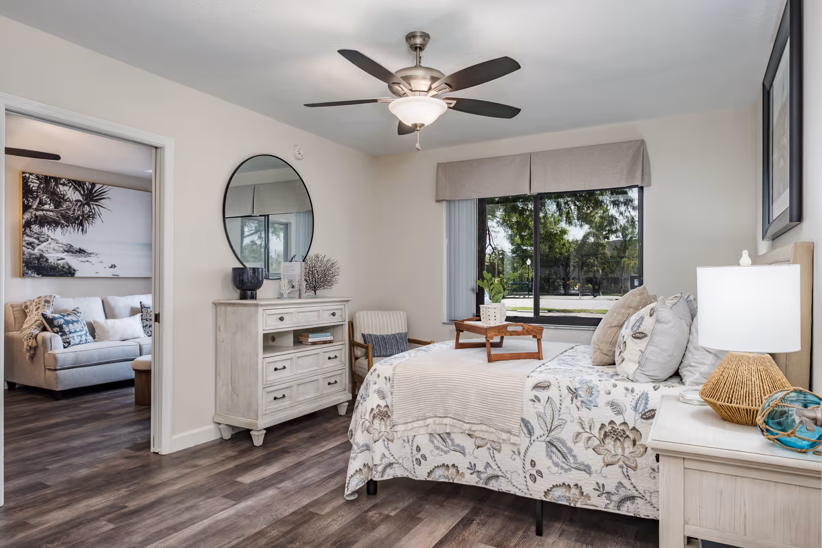A bright bedroom with a floral patterned bedspread, multiple pillows, a wooden tray on the bed, a bedside table with a lamp, and a dresser with a round mirror above it. A window with a view of trees and a street is on one wall. Through an open doorway, a living room with a sofa and large wall art is visible.