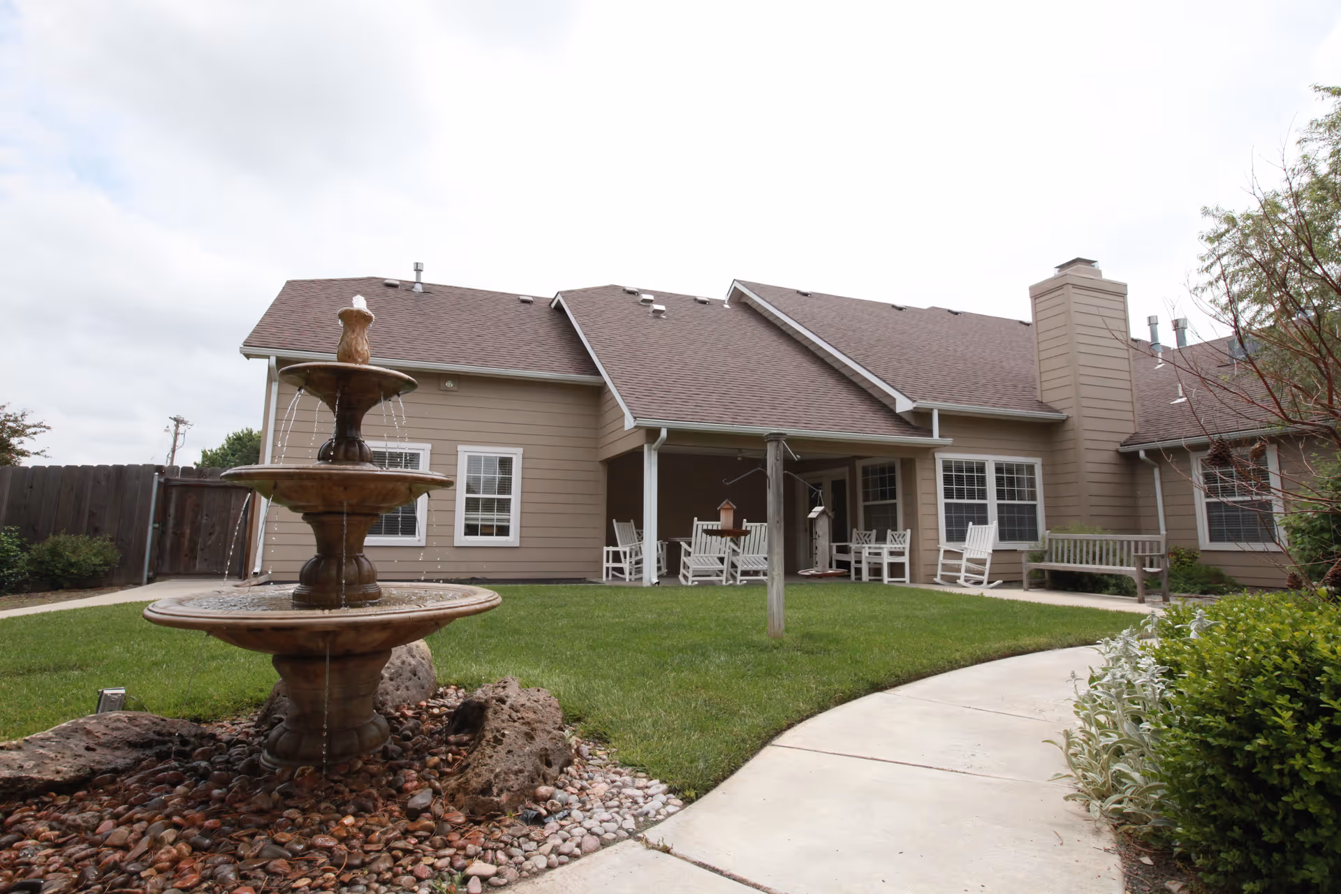 Outdoor courtyard area of a senior living facility featuring a three-tiered water fountain surrounded by rocks, a curved concrete pathway, green grass, and a covered patio with white rocking chairs and benches. The building has beige siding and multiple windows under a brown shingled roof.