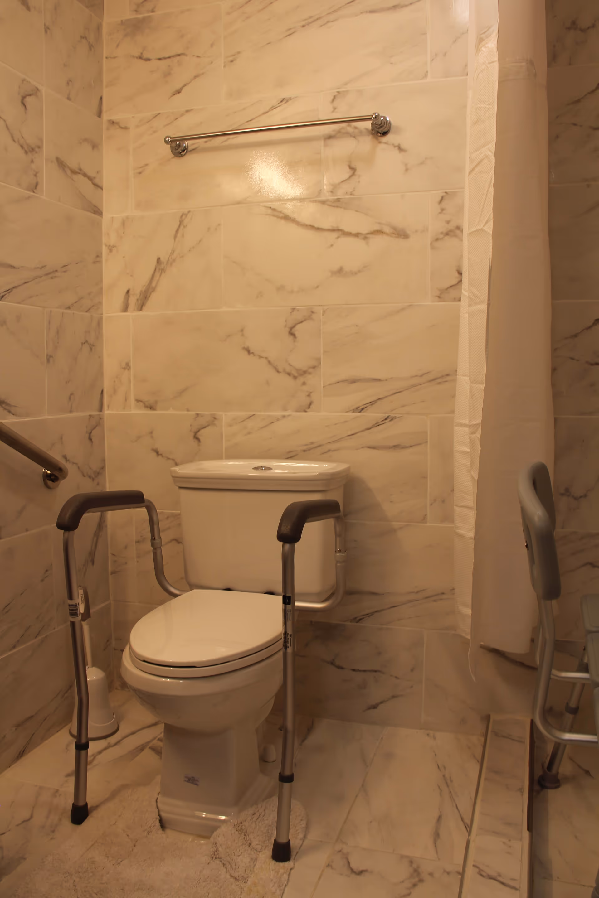 A bathroom with marble-patterned tile walls and floor, featuring a white toilet with safety rails on both sides, a white toilet brush holder, a towel bar above the toilet, and a shower area with a white curtain and a shower chair.