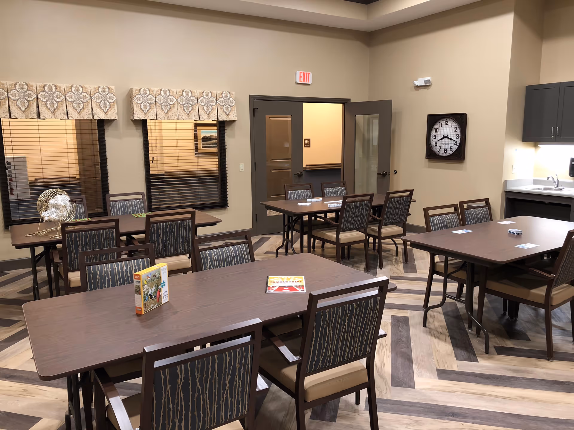 A well-lit room with several rectangular tables and chairs arranged for group activities or dining. The room has patterned valances over two windows with blinds, a wall clock, and a kitchenette area with a sink and cabinets. There are game cards and a bingo cage on the tables, suggesting a recreational or communal space.