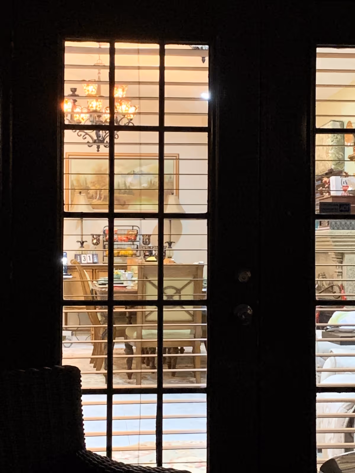 View through glass-paneled doors with horizontal blinds partially open, showing a warmly lit dining room with a chandelier, a dining table surrounded by chairs, a sideboard with decorative items, and a framed painting on the wall.