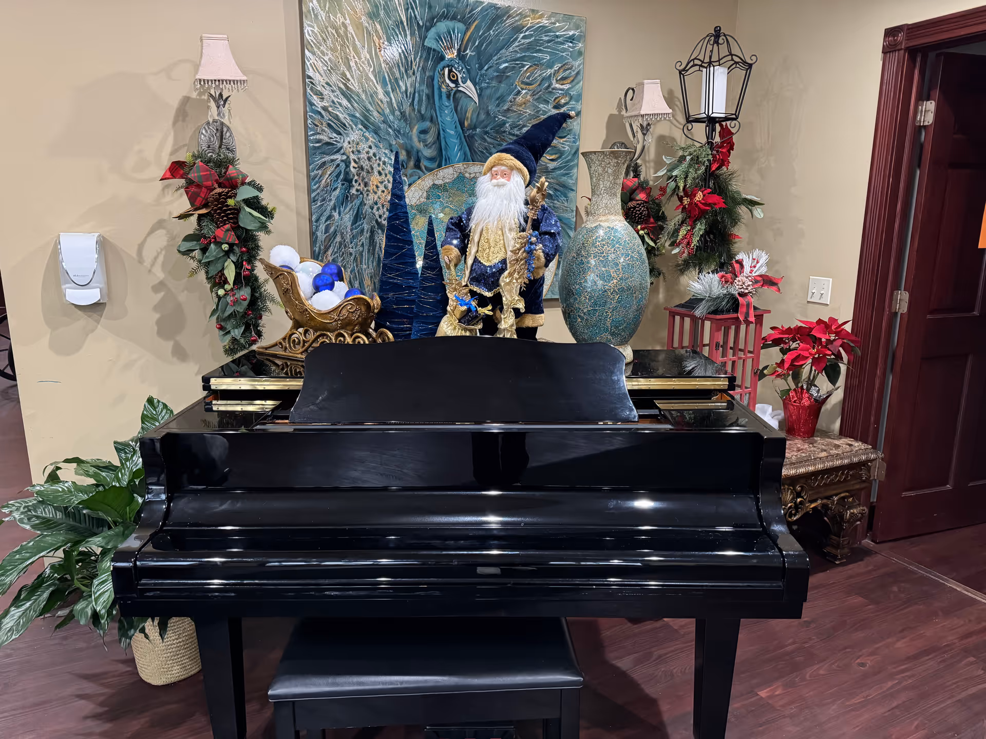 A black grand piano with a matching bench in front of a beige wall. On top of the piano are decorative items including a Santa Claus figurine dressed in blue and gold, a large blue and gold vase, a sleigh with blue and white ornaments, and two tall blue cone-shaped decorations. Behind the piano is a large painting of a peacock with blue and green feathers. The wall is decorated with two wall lamps adorned with holiday greenery and red ribbons. To the right, there is a small table with a red poinsettia plant and holiday decorations. A dark wooden door is visible on the right side of the image.