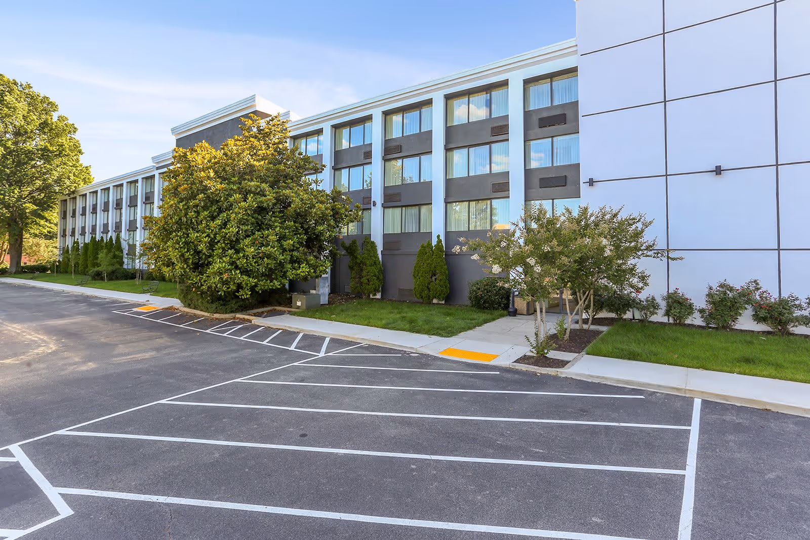 Exterior view of a multi-story senior living facility building with large windows, surrounded by green trees and shrubs, and an empty parking lot in the foreground.