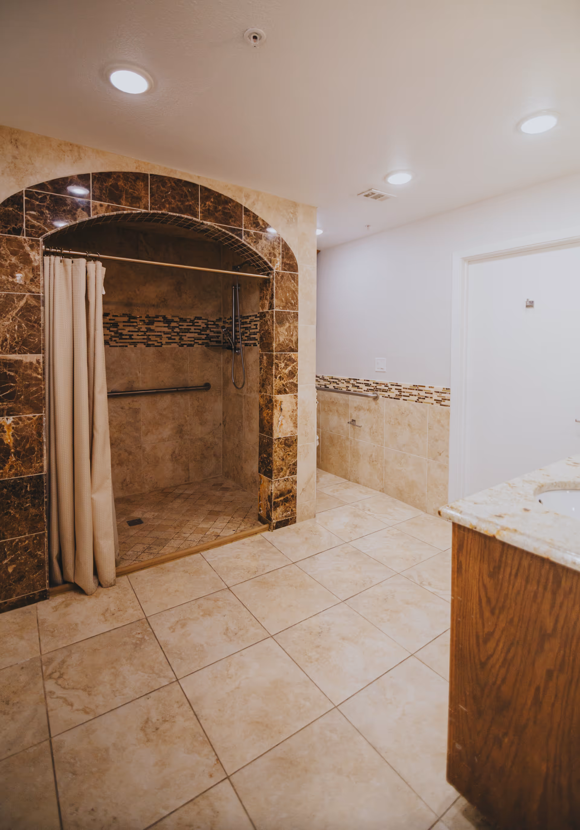 A bathroom with beige tiled floors and walls, featuring a walk-in shower with a curtain and dark brown tile archway. The shower has a grab bar and a handheld showerhead. There is a wooden vanity with a granite countertop visible on the right side.