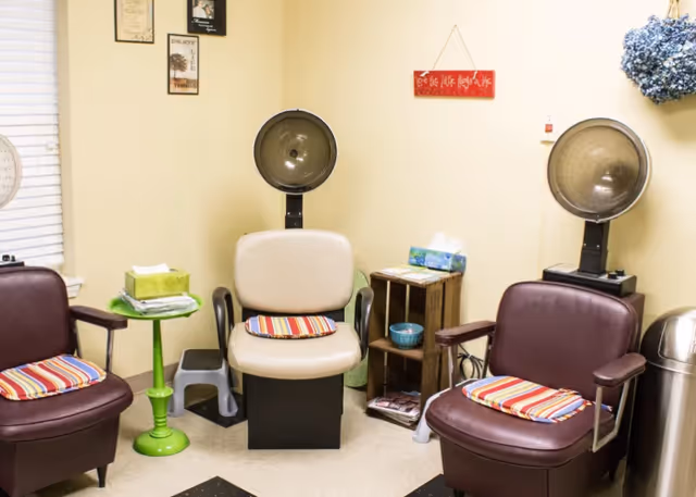 A small salon area with three vintage hair dryer chairs, each with a striped cushion. The walls are light yellow with framed pictures and a red sign that reads 'Love the little things in life.' There is a green side table with tissues and towels, a small wooden shelf with magazines and tissues, and a silver trash can.