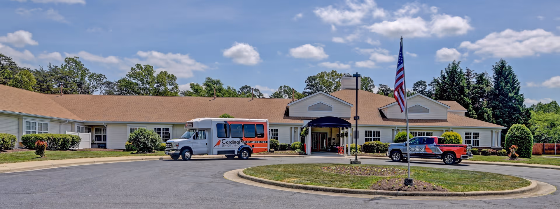 Exterior front view of Cardinal Senior Living at Danville, showing a single-story building with a brown roof and white walls. There is a circular driveway with a flagpole in the center, an American flag flying, and two vehicles parked near the entrance, one of which is a shuttle bus with the Cardinal logo. Trees and bushes surround the building under a partly cloudy sky.