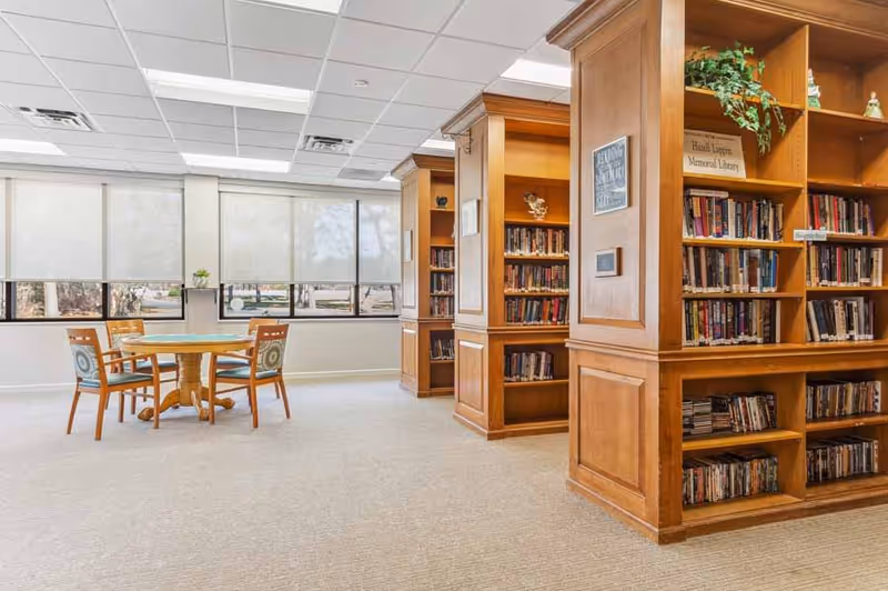 Bright communal library with wooden bookshelves and a round table surrounded by chairs near large windows.