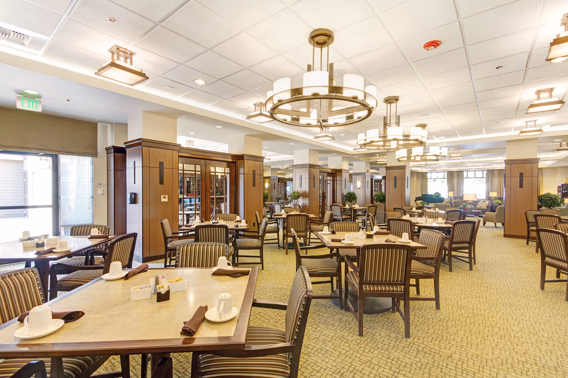 A spacious dining room with multiple tables and chairs arranged neatly. Each table is set with white cups, saucers, and brown napkins. The room features large circular chandeliers hanging from a white ceiling with recessed lighting. The walls have wooden paneling and there are large windows allowing natural light to brighten the space. The overall atmosphere is warm and inviting.