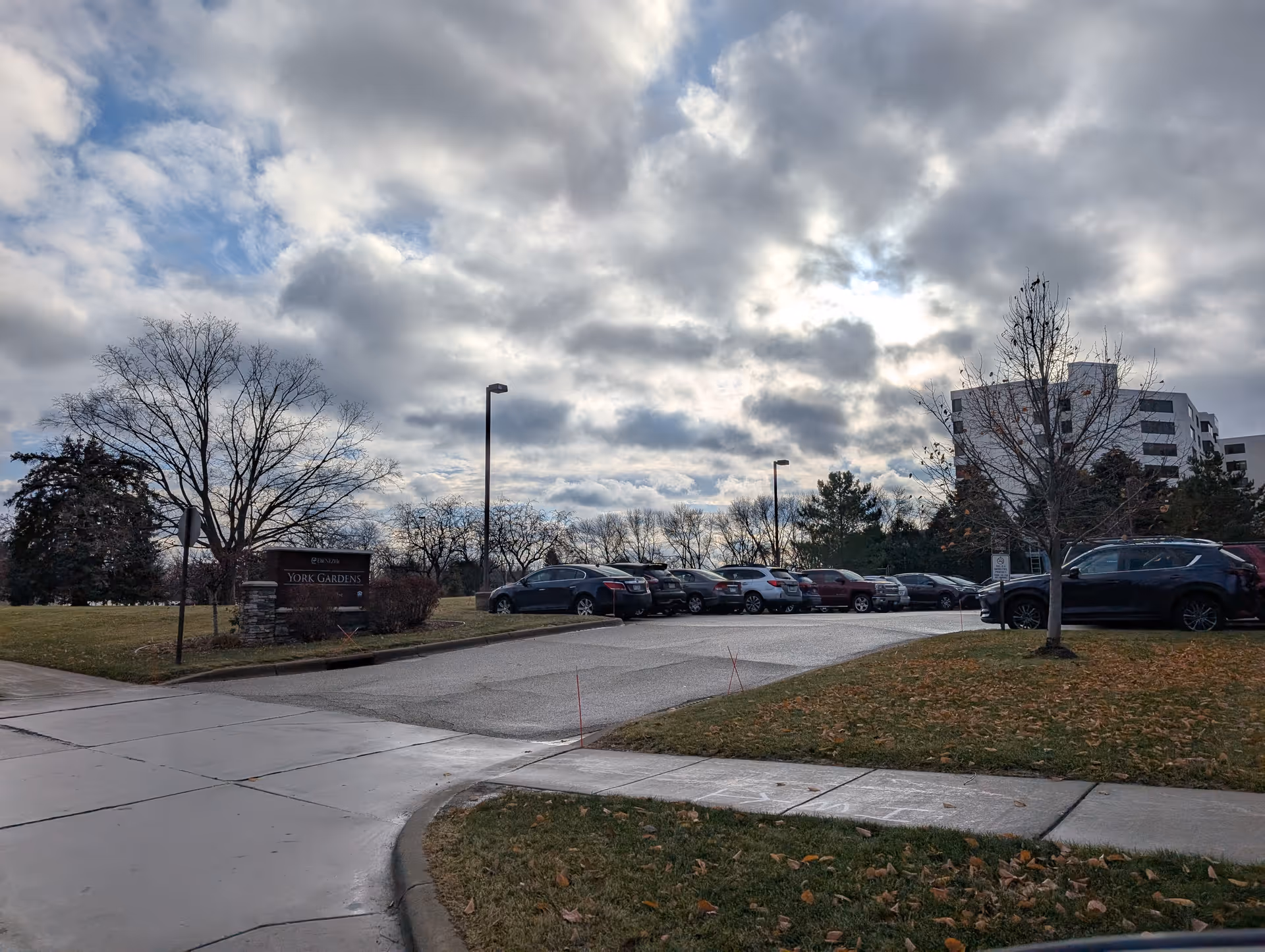 Parking lot area outside York Gardens facility with several parked cars, leafless trees, a cloudy sky, and a sign that reads 'York Gardens'.