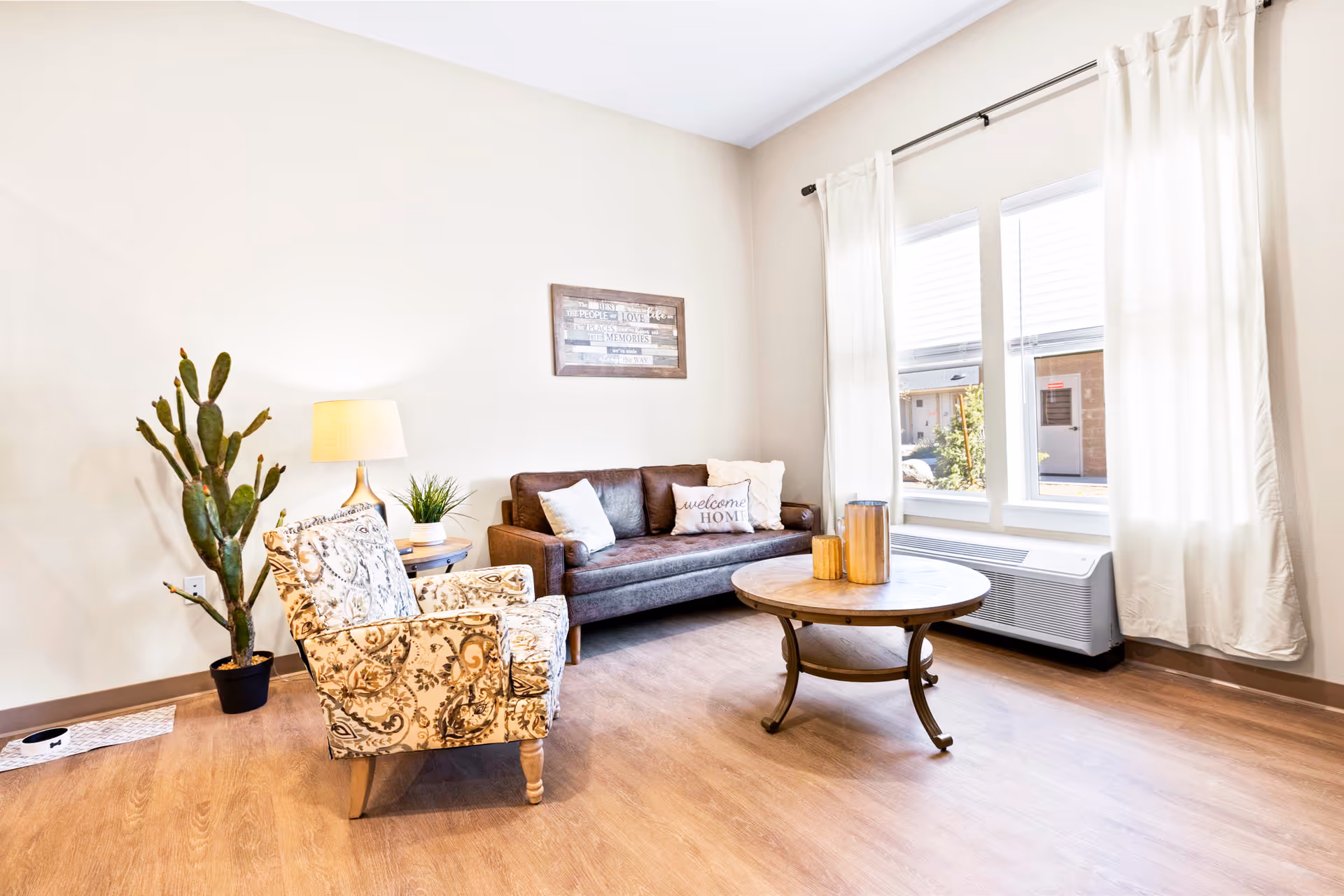 Bright living room featuring a sofa, patterned armchair, round wooden coffee table, potted cactus and a large window with curtains.