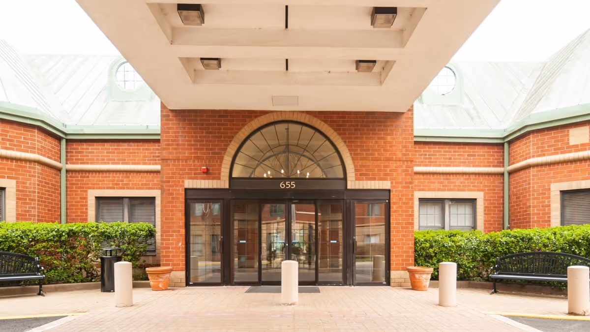 Entrance of a brick building with large glass double doors under an arched window. The building number 655 is displayed above the doors. There are green bushes, two black benches, and two large terracotta pots on either side of the entrance. The area is covered by a white overhang with recessed lighting.