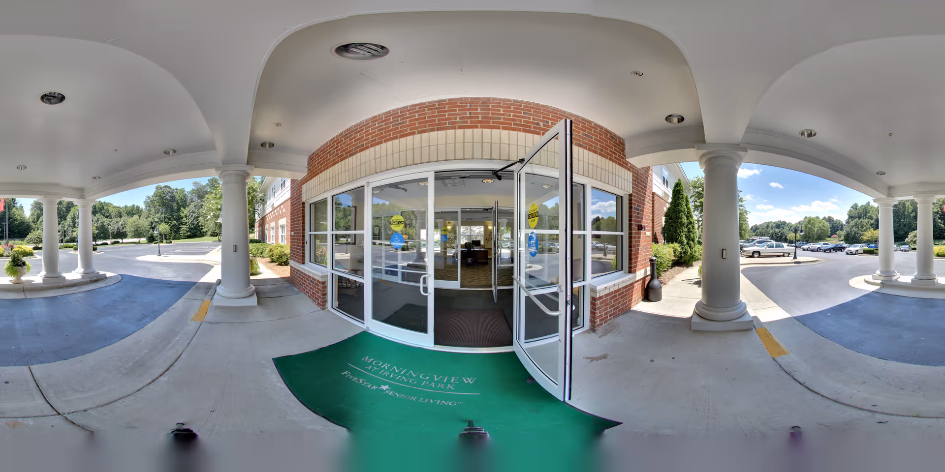 Entrance to Morningview at Irving Park senior living facility showing a covered driveway with white columns, glass double doors, and a green welcome mat with the facility's name. The surrounding area includes a parking lot and greenery under a clear blue sky.