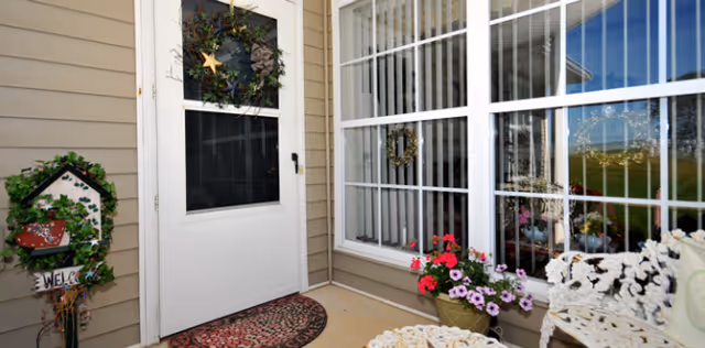 Front porch area of a residential unit with a white door decorated with a wreath, a welcome sign with greenery on the left, a window with white bars and a wreath inside, a flower pot with pink and purple flowers, and white metal outdoor furniture.