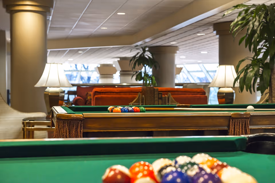 Interior view of a recreational room with two pool tables in the foreground and background. The room features large columns, table lamps, plants, and red sofas with large windows letting in natural light.