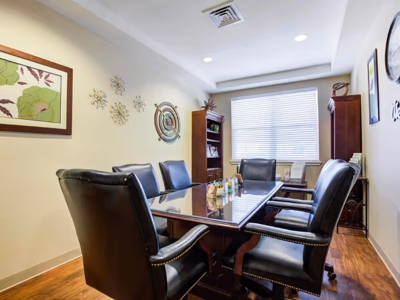 A small conference room with a rectangular wooden table surrounded by six black leather chairs. The room has light-colored walls decorated with framed artwork and metal wall sculptures. There is a window with blinds letting in natural light, and wooden bookshelves with decorative items and books.