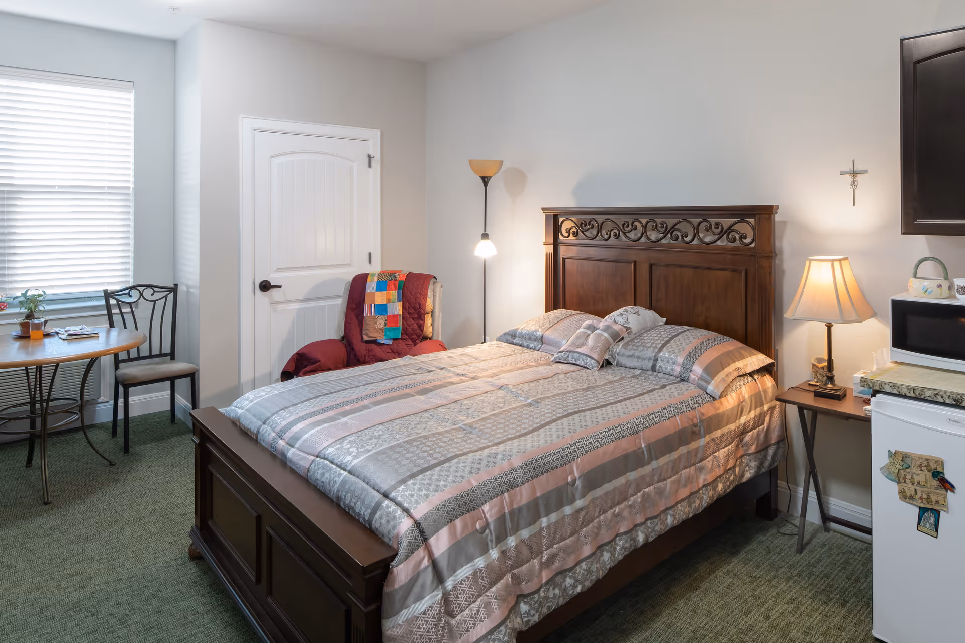 A cozy bedroom in an assisted living facility featuring a wooden bed with a patterned bedspread and pillows, a red armchair with a colorful quilt draped over it, a small round table with two chairs near a window with blinds, a floor lamp, a bedside table with a lamp, a microwave on a small counter, and a mini refrigerator with magnets. A cross hangs on the wall above the bedside table.