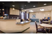 Well-lit dining area with round tables, chairs, and a service counter by kitchenette.