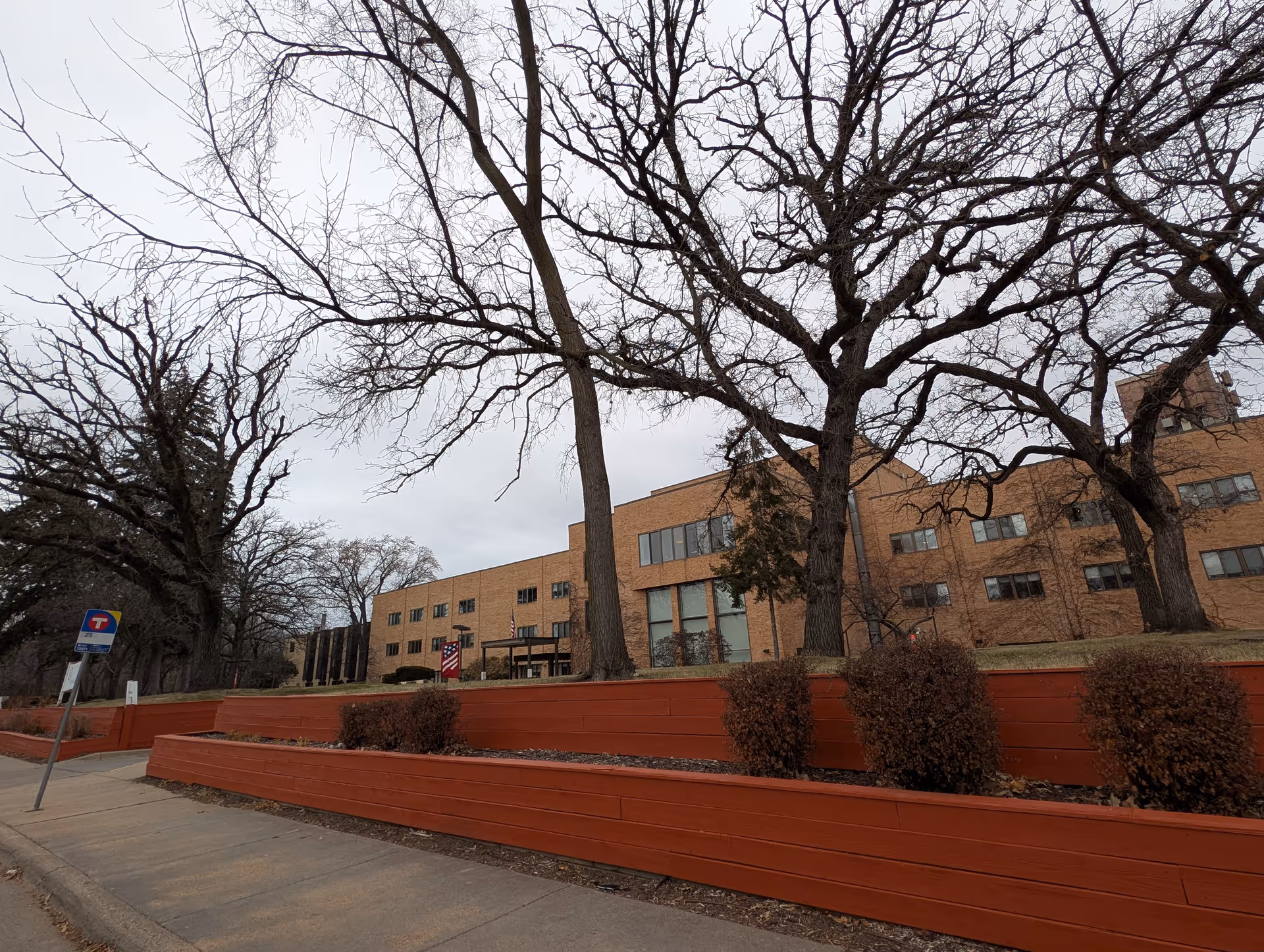 Exterior view of a large brick building with multiple windows, surrounded by leafless trees and red wooden retaining walls along the sidewalk under an overcast sky.