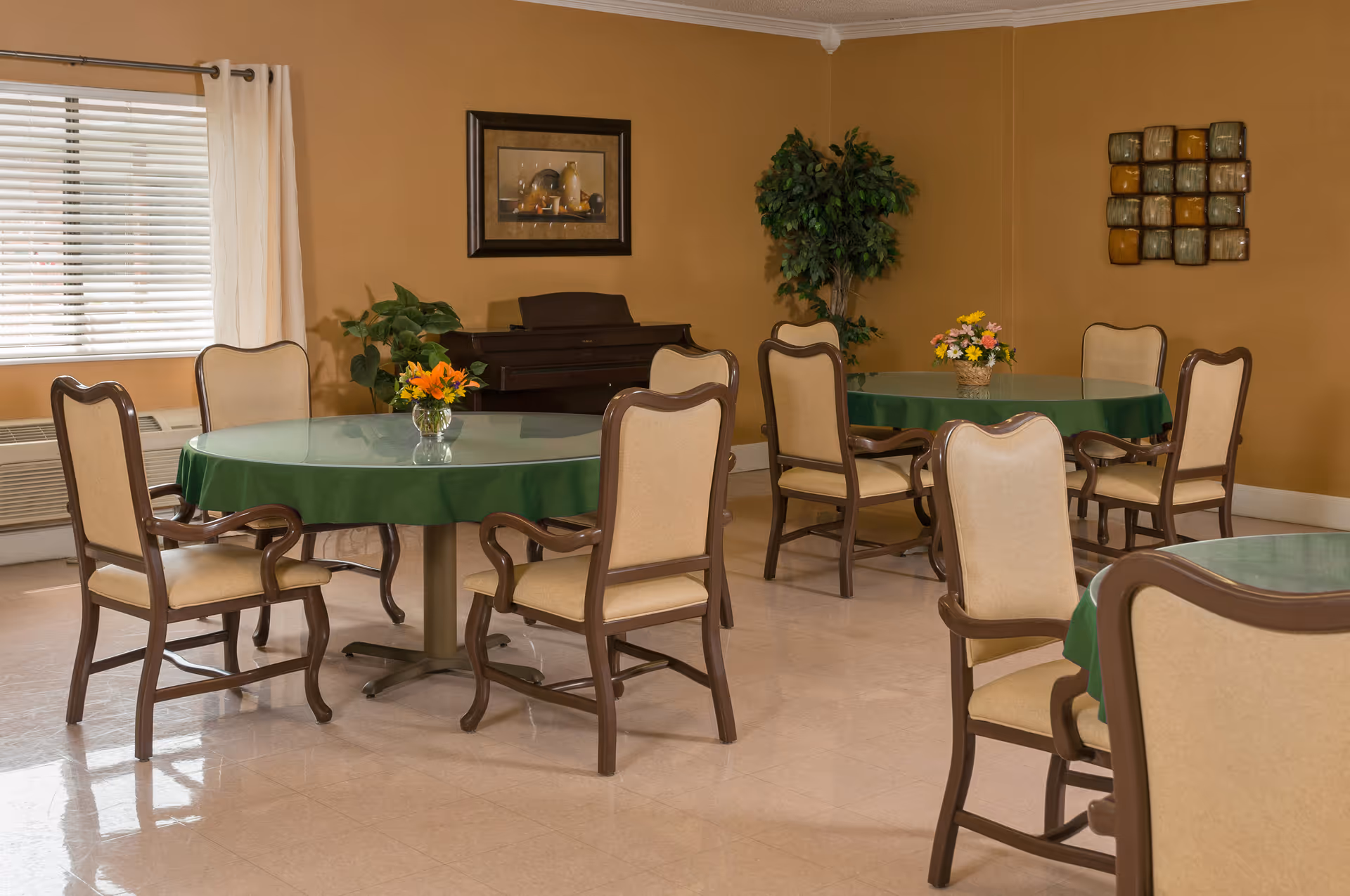 A dining room with round tables covered in green tablecloths and glass tops, surrounded by beige cushioned chairs with wooden frames. There are flower arrangements on the tables, a window with white curtains, a piano against the wall, a potted plant, and framed artwork on the tan-colored walls.
