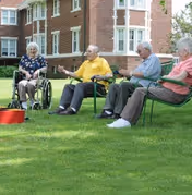 Four elderly individuals sitting outdoors on a grassy lawn near a brick building. Two are seated in wheelchairs, and two are seated on green metal chairs. They appear to be engaged in conversation or a group activity on a sunny day.