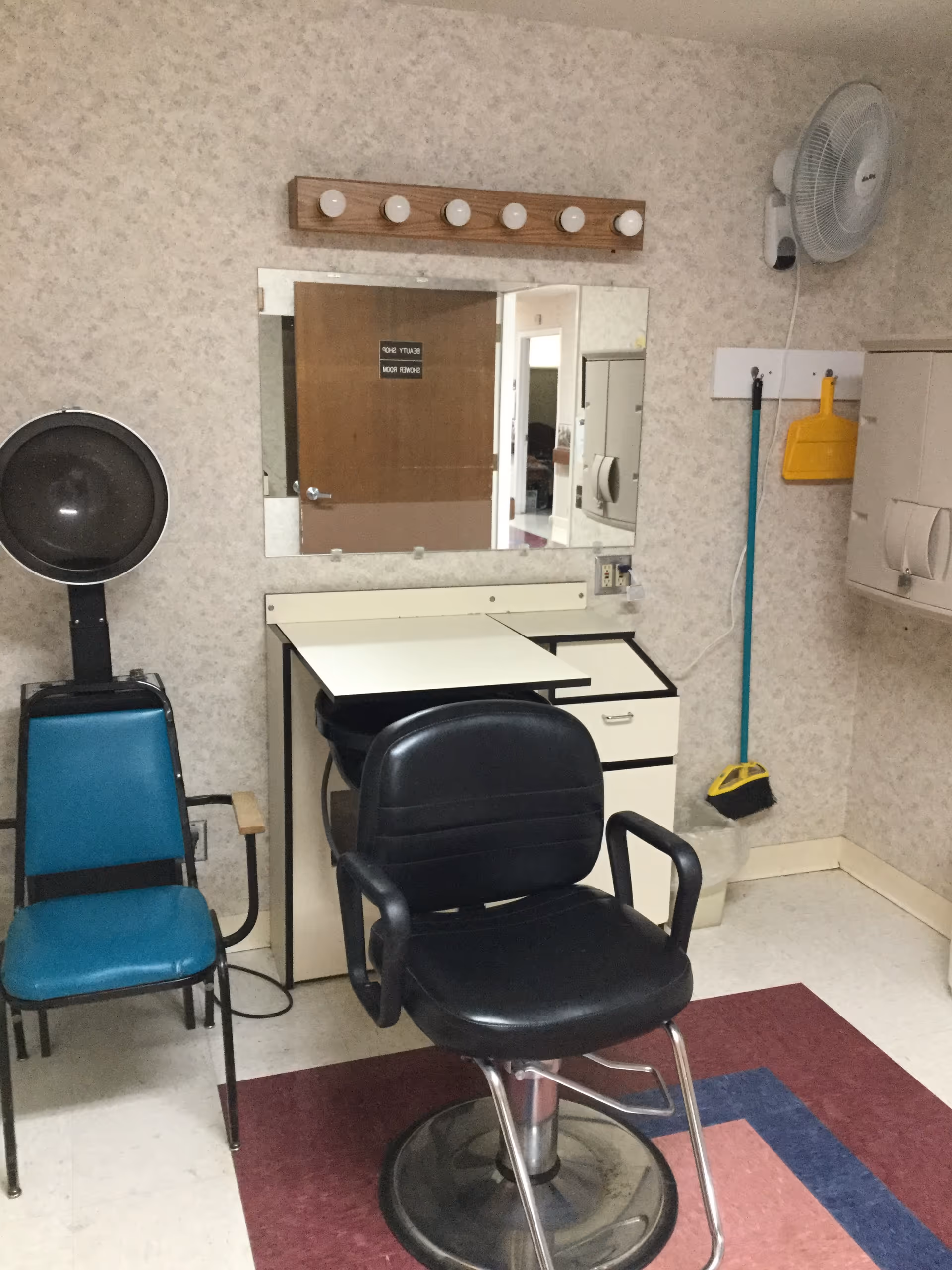 A small salon or beauty room with a black salon chair in front of a vanity mirror with lights above it. To the left is a blue chair with a hair dryer hood attached. On the right wall, there is a mounted fan, a broom, and a dustpan hanging on hooks. The floor has a red and blue patterned rug.