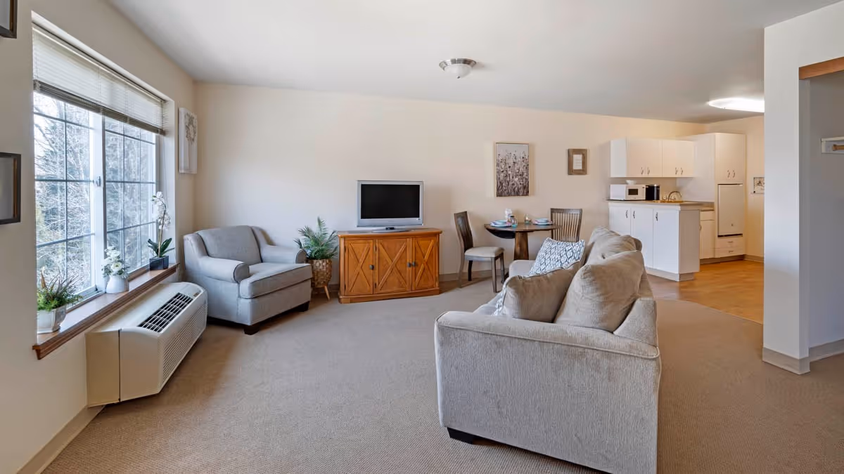 A bright and spacious living area in a senior living facility with a large window on the left, a gray armchair, a wooden TV stand with a flat-screen TV, a small round dining table with two chairs, and a beige sofa facing away from the camera. The room opens into a kitchen area with white cabinets and appliances.