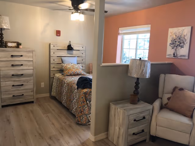 A cozy bedroom in an assisted living facility featuring a single bed with patterned bedding, a wooden headboard, and a matching dresser. Next to the bed is a small nightstand with a lamp. A comfortable armchair with a pillow is positioned near a window with blinds, and the walls are painted in soft beige and peach tones.